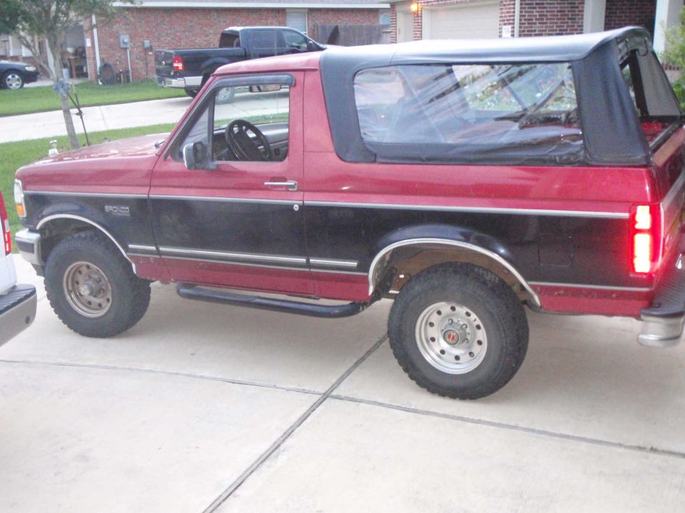 Side view of a red and black 1994 Ford Bronco with a black soft top. A black pickup truck is parked in the background
