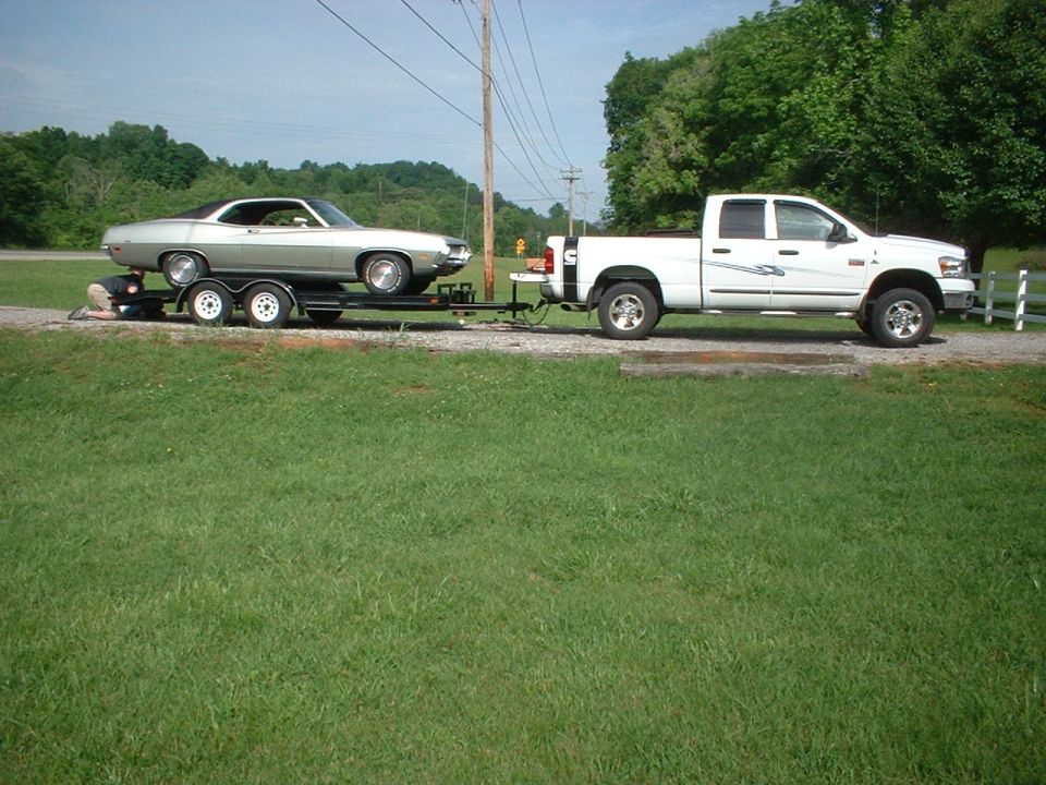 Silver coupe on a trailer, towed by a white pickup truck. Side view