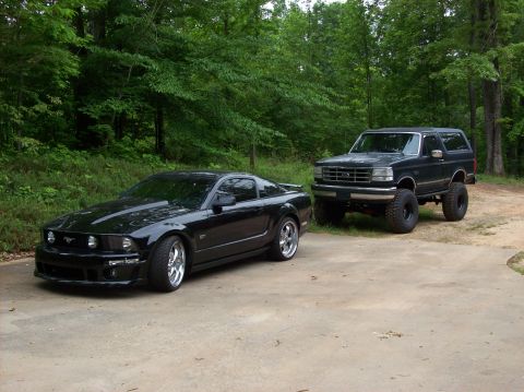 2007 Ford Mustang - Two vehicles, a modern Mustang and an older lifted Bronco, are parked on a concrete driveway with trees in the background.