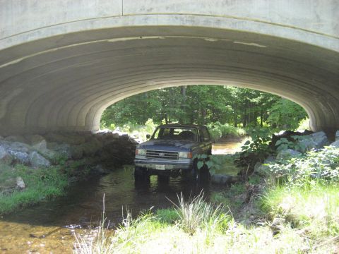 1991 Ford Bronco - Ford Bronco navigates a shallow creek beneath a concrete overpass on a wooded trail.