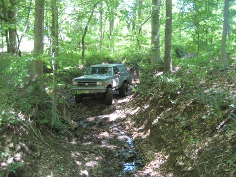 1991 Ford Bronco - A green 1991 Ford Bronco navigates a challenging off-road path through a wooded area.