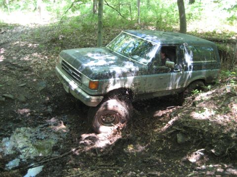 1991 Ford Bronco - A Ford Bronco navigates a challenging muddy terrain in a wooded area.