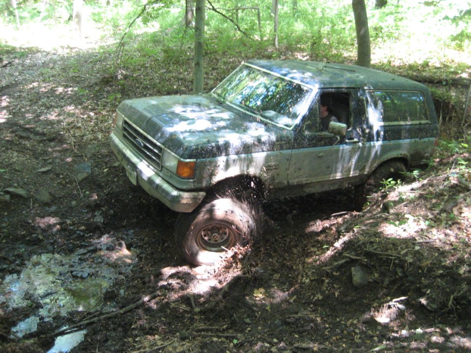 Mud-splattered dark gray and silver Ford Bronco stuck in a muddy forest trail