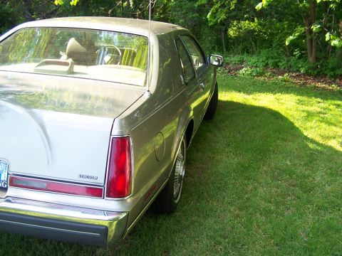 1987 Lincoln Mark VII - The rear of a silver Lincoln Mark VII is visible, parked on a grassy lawn with trees in the background.