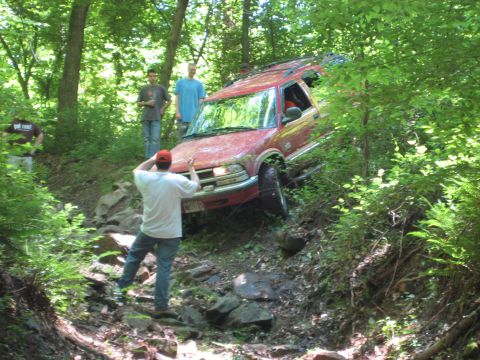 1991 Ford Bronco - A red SUV is being driven off-road on a challenging, rocky path through a wooded area.