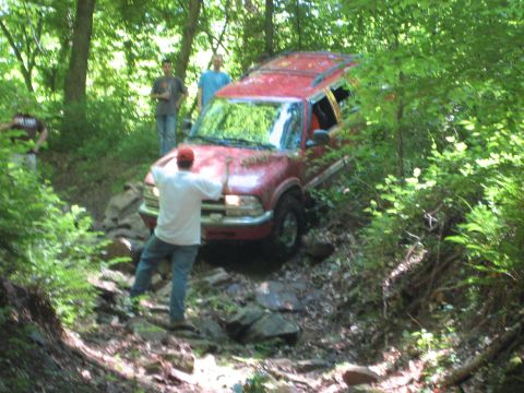 1991 Ford Bronco - A red SUV is being driven off-road through a wooded area with several people observing.