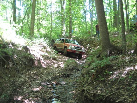 1991 Ford Bronco - An orange SUV navigates a challenging off-road path through a dense forest.
