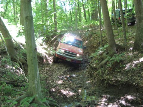 1991 Ford Bronco - A red SUV tackles a challenging muddy trail in a wooded area, with another vehicle in the distance.