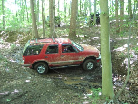 1991 Ford Bronco - A red SUV navigates a muddy trail in a wooded area, with other vehicles visible in the distance.