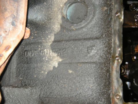 1979 Ford Bronco - Close-up of an engine block casting showing stamped markings and a section of rusty exhaust pipe.