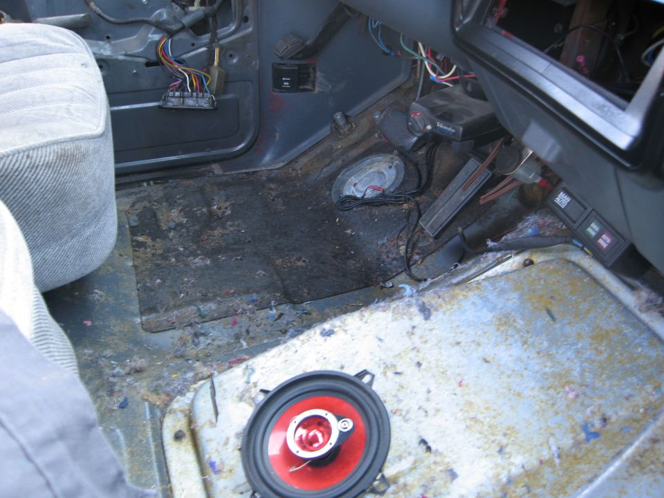 Interior floor of a vehicle showing exposed wiring, a removed carpet section, and a loose speaker