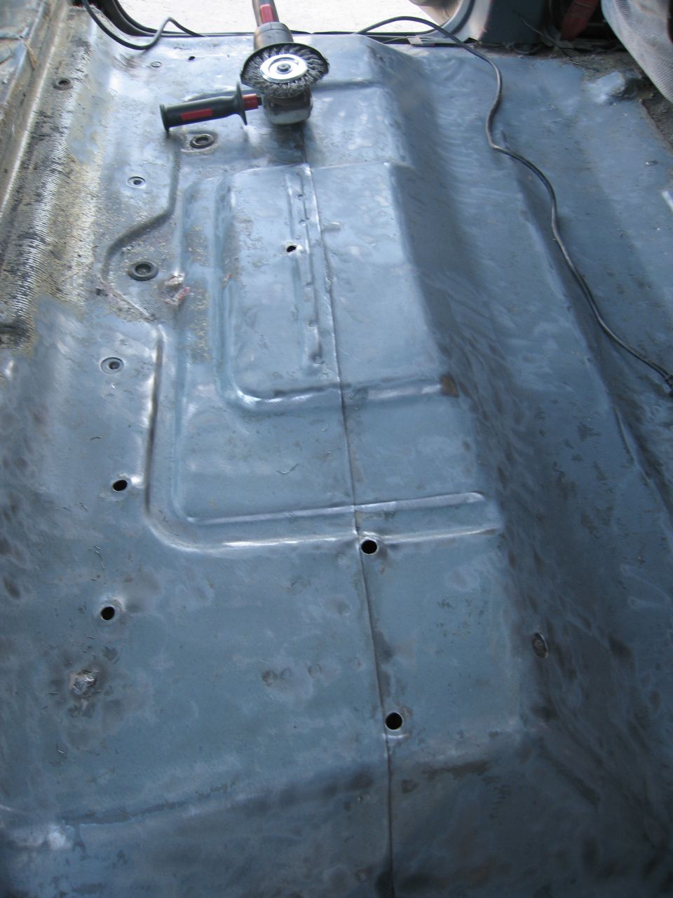 Bare metal vehicle floor with a grinder tool on it, showing rust and repair welds