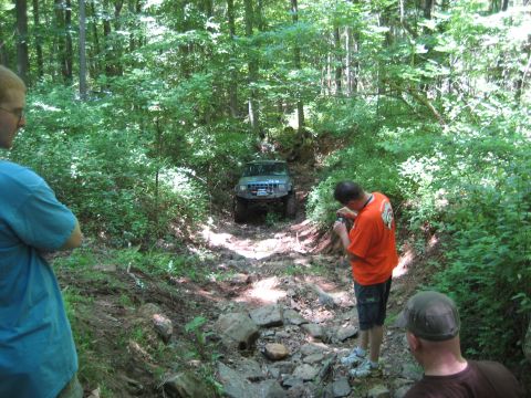 1991 Ford Bronco - Two SUVs tackle a challenging, rocky trail surrounded by dense forest foliage.