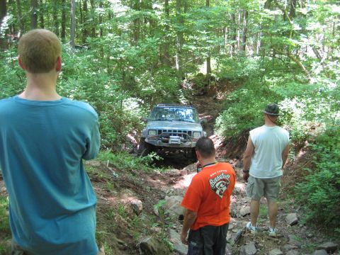 1991 Ford Bronco - A Jeep Cherokee XJ navigates a rocky, wooded path with onlookers present.