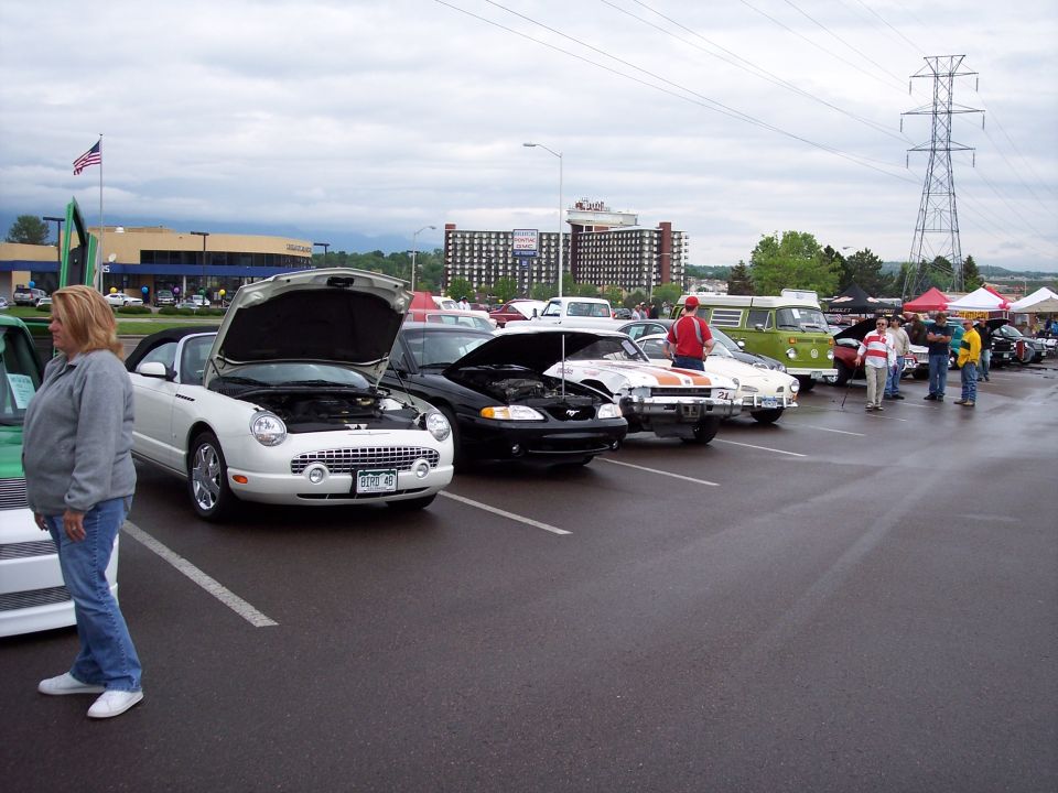 Several cars parked in a lot with hoods open, people milling around, and tents in the background