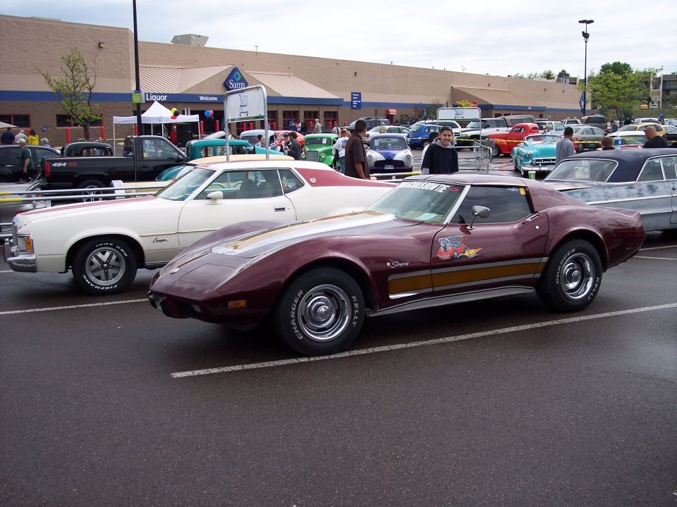 Maroon 1975 Chevrolet Corvette C3 with gold stripes and racing decals parked next to a white Ford coupe