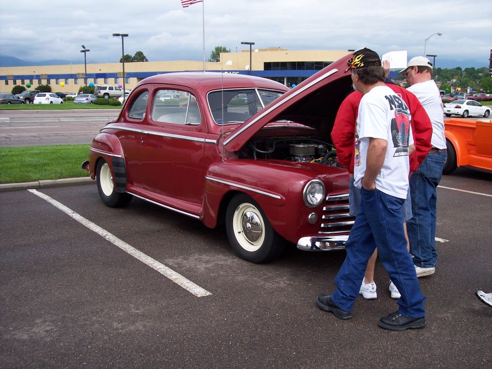 Red coupe with hood open, white wall tires, chrome grille. Three men looking at engine