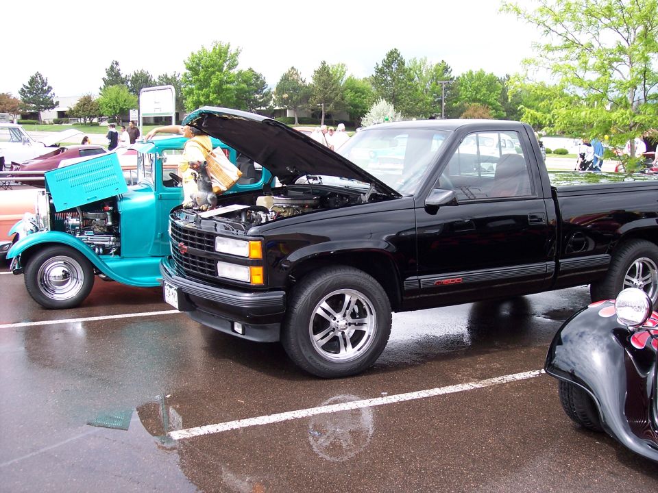 Black pickup truck, hood open, parked next to a teal hot rod