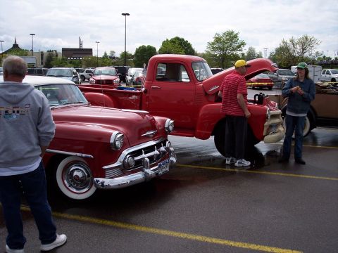 1972 Ford Ranchero - Two classic red vehicles, a sedan and a pickup truck, are parked on a wet asphalt lot.