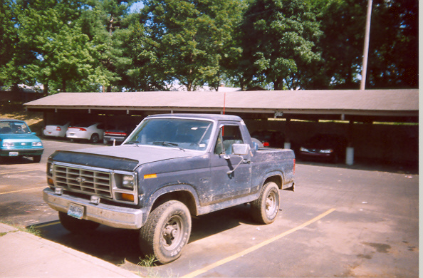 Blue Ford pickup-style Bronco parked under a carport row of trees, front-quarter view