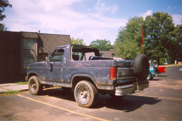 Dark blue Ford Bronco, top off with tailgate-mounted spare, parked outside a building