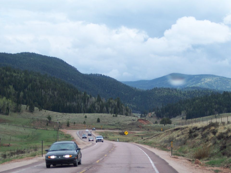 Several cars drive on a highway winding through a valley with forested mountains under a cloudy sky