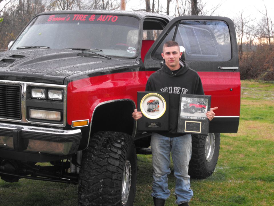 Red and black lifted pickup truck with a man holding awards. The award on the right shows "1987 1997"