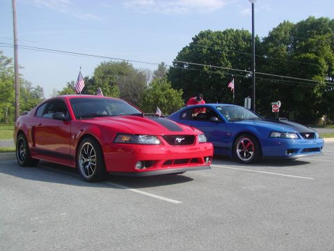 2004 Ford Mustang - Two Ford Mustangs, one red and one blue, are parked side-by-side in a lot, both racing stripes and American flags.