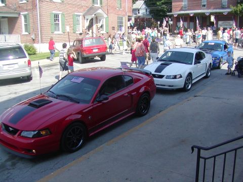 2004 Ford Mustang - A red 2004 Ford Mustang is parked on the street, with other vehicles and spectators in the background.