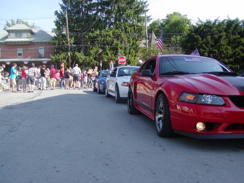 2004 Ford Mustang - A red 2004 Ford Mustang leads a line of cars in a parade, with spectators lining the street.
