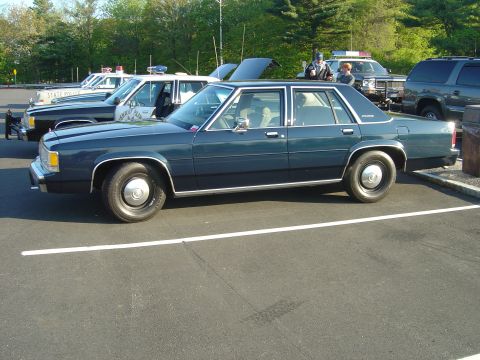 2001 Ford Crown Victoria - A dark blue sedan is parked in a lot with several police vehicles visible in the background.