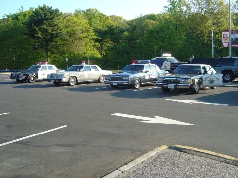 2001 Ford Crown Victoria - A lineup of four police vehicles, including a Highway Patrol car with its door ajar, are parked in a lot on a sunny day.