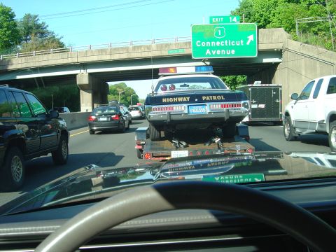 2001 Ford Crown Victoria - A police car is being towed on a trailer on a busy highway under an overpass.