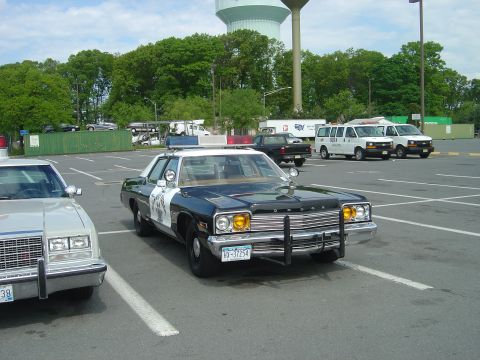 2001 Ford Crown Victoria - This 1977 Dodge Monaco police car is parked in a lot with other vehicles and trees in the background.