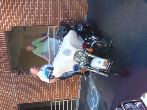 2001 Ford Crown Victoria - Two men stand next to a white police motorcycle parked in front of a brick building.