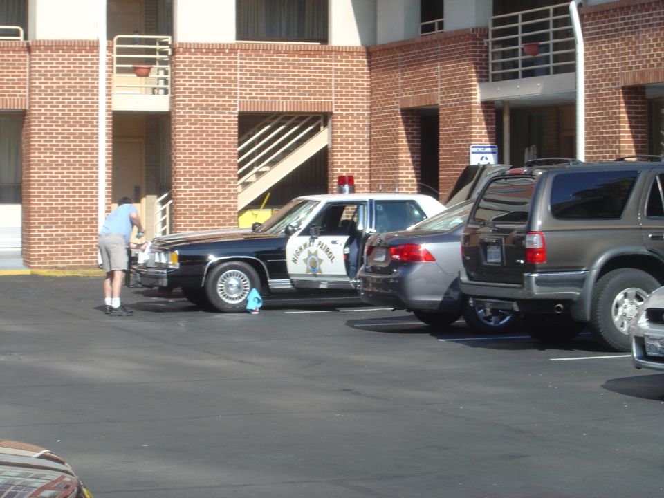Police car with 'Highway Patrol' on door being washed by a man in shorts and blue shirt. Other cars parked nearby