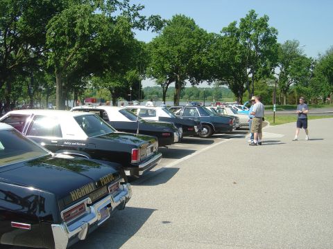 2001 Ford Crown Victoria - A line of older model police cars are parked in a lot on a sunny day, with trees and a body of water in the background.