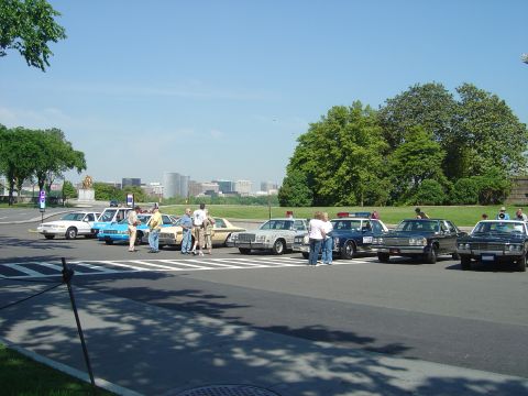 2001 Ford Crown Victoria - A collection of vehicles, including several police cruisers, are gathered outdoors on a clear day.