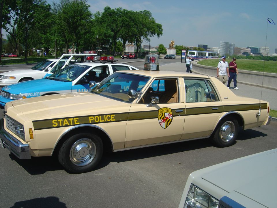 Tan 1977 police car with 'STATE POLICE' text and Maryland seal on the door, parked next to a blue and white police car
