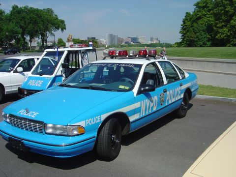 2001 Ford Crown Victoria - A blue and white NYPD police car is parked on asphalt, with a white police van and another vehicle nearby under a clear sky.