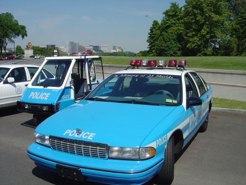 2001 Ford Crown Victoria - Two police vehicles, one a sedan and the other a three-wheeled unit, are parked outdoors with a city skyline in the background.