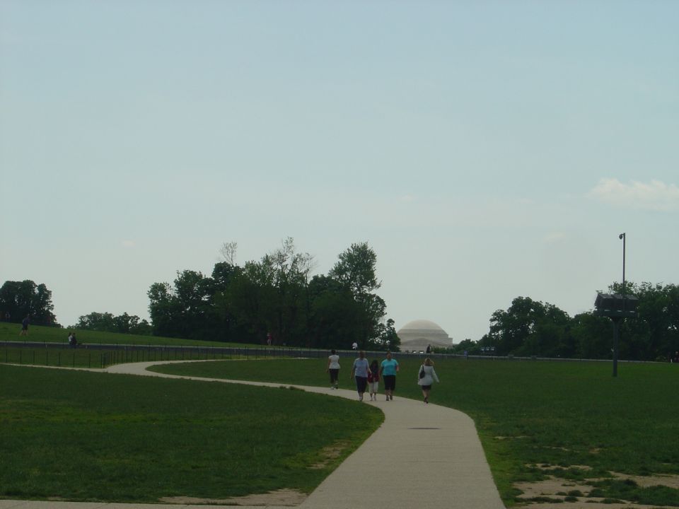 People walk on a paved path through a grassy park towards a distant domed building