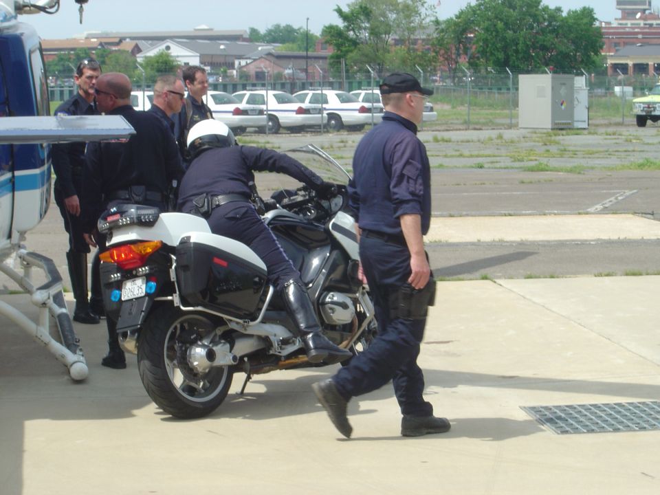 Police officers gather near a motorcycle and helicopter, with several white sedans parked in the background