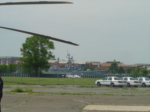 2001 Ford Crown Victoria - A line of police cruisers are parked in an open lot, with a large naval vessel visible behind a fence.