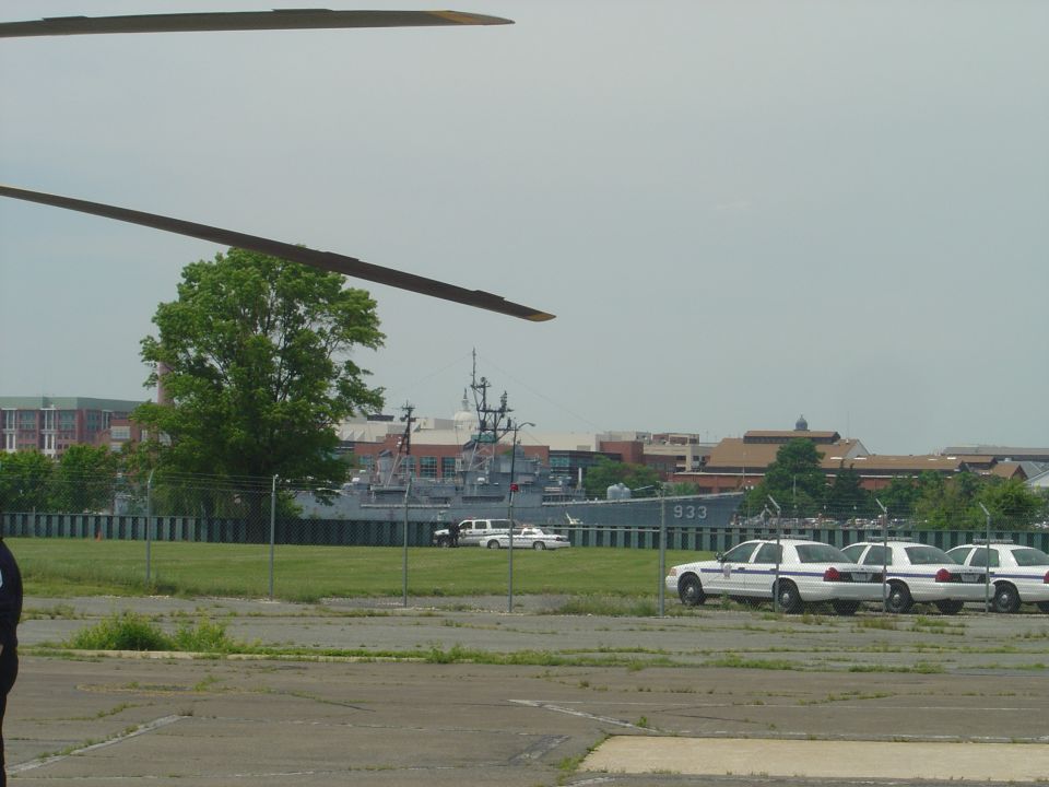 Several white police cars parked in a lot with a naval ship in the background