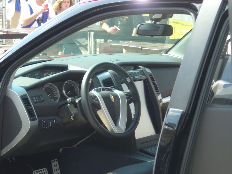 Car interior with a large screen and a steering wheel with a diamond logo. Text on screen reads "CARBON MOTORS"