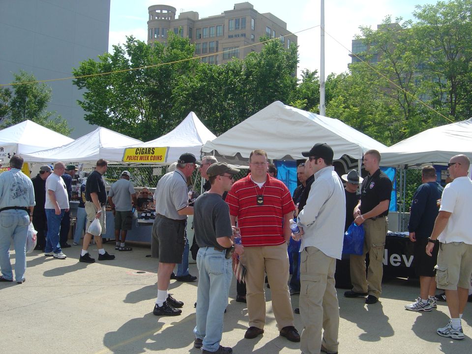 Outdoor event with tents and people, "CIGARS POLICE WEEK COINS" sign visible