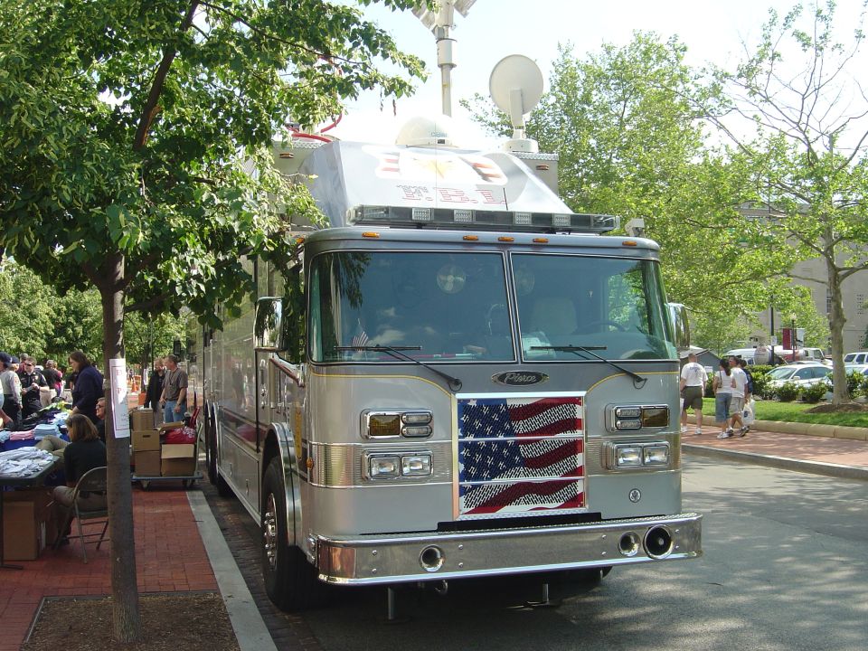 Front view of a silver fire truck with an American flag decal on the grille. People and trees are visible in the background