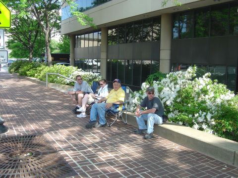 2001 Ford Crown Victoria - Four men relax on a sunny day, seated on folding chairs along a brick walkway next to a building and blooming shrubs.
