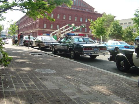 2001 Ford Crown Victoria - Several police vehicles are parked along a street in front of a large brick building.